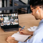 A male office employee, takes notes during online video conference with mature successful ceo and multiracial colleagues, using laptop, sitting in office, planning marketing strategy