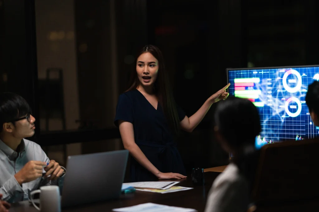 A professional woman presents data on a screen to colleagues in a conference room.