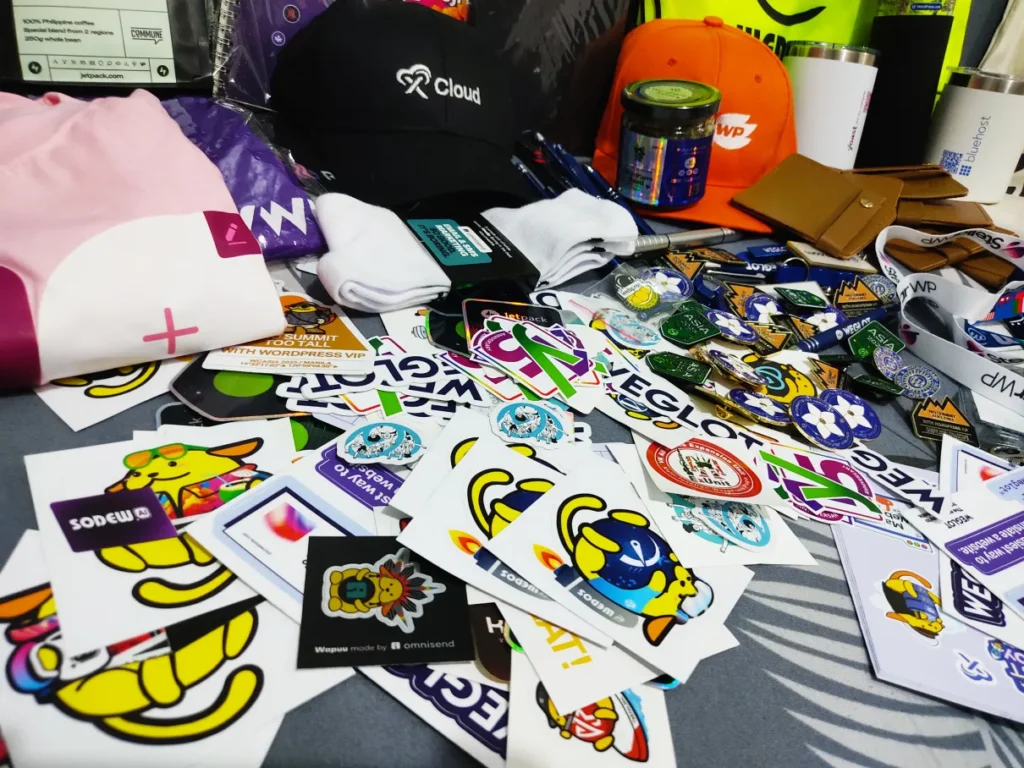 A tabletop cluttered with an assortment of colorful stickers, promotional items, and merchandise from WordCamp Asia 2025, showcasing the event's community spirit