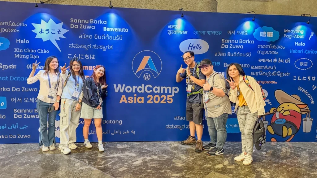 Members of the Web Development Team posing in front of a colorful wall covered with greetings in various languages at WordCamp Asia 2025