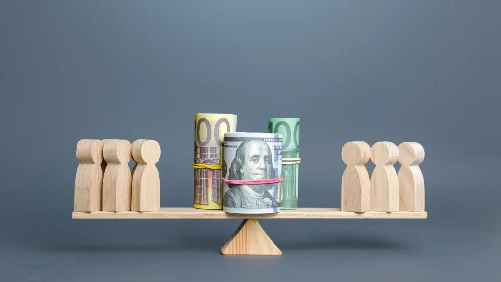 A wooden balance scale with money on one side and small figures representing people on the other side.