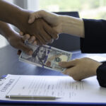 Two people shaking hands while exchanging cash over a signed contract on a desk.