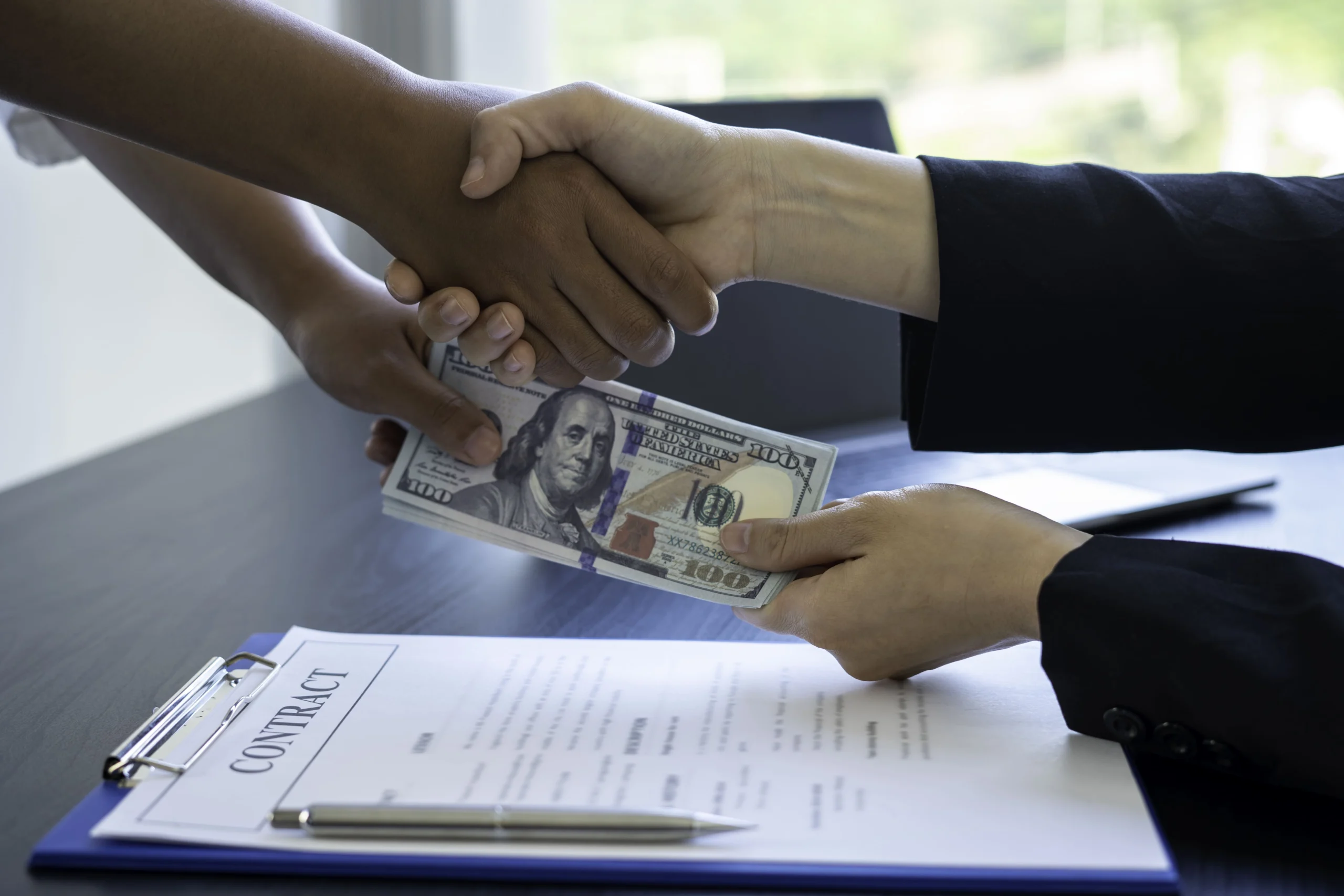 Two people shaking hands while exchanging cash over a signed contract on a desk.