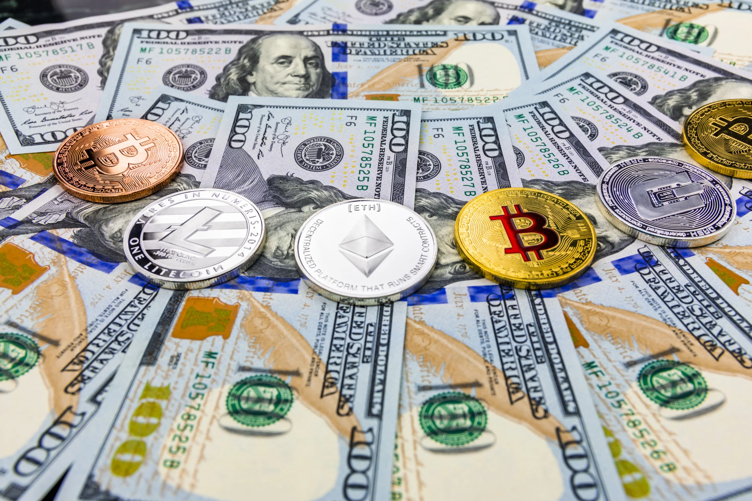 Assorted physical cryptocurrency coins including Bitcoin, Ethereum, Litecoin, and others displayed on top of scattered US $100 bills.
