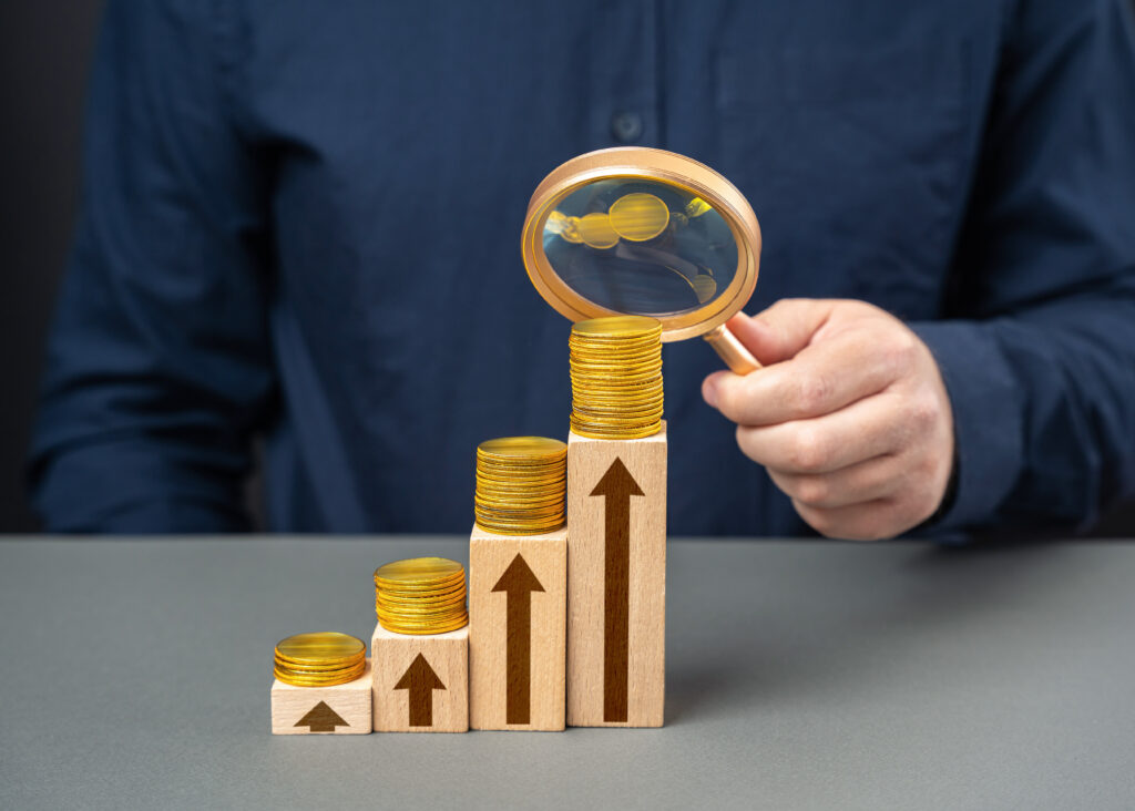 A businessman analyzing financial growth with a magnifying glass over stacked coins on ascending wooden blocks, each marked with upward arrows, symbolizing increasing income or investment returns
