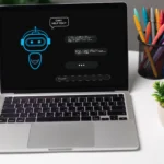 A laptop on a desk shows a chatbot interface on its screen, with a speech bubble saying "Can I help you?" Nearby are colorful pens, a small plant, and a coffee cup, creating a workspace vibe.