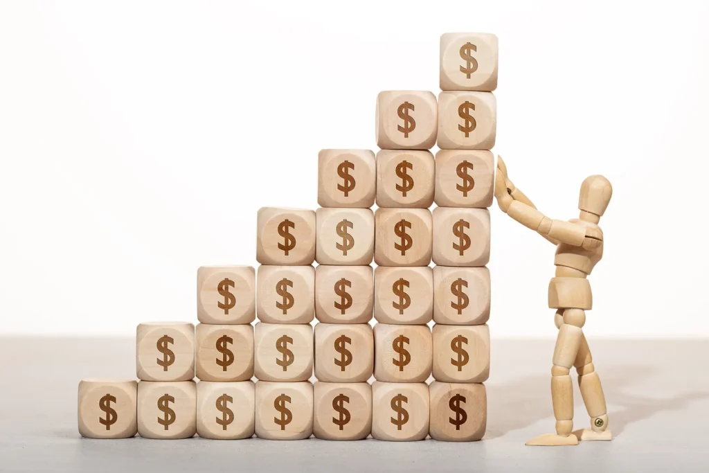 A wooden figure stands atop a stack of wooden blocks, each marked with dollar signs, symbolizing financial success.