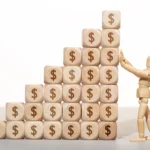 A wooden figure stands atop a stack of wooden blocks, each marked with dollar signs, symbolizing financial success.