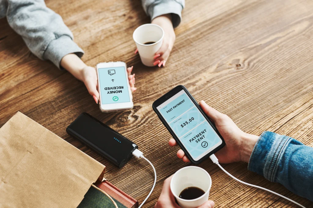 Two people sitting at a wooden table exchanging money via mobile phones. Both hold coffee cups, creating a casual and modern tech-savvy ambiance.