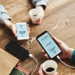 Two people sitting at a wooden table exchanging money via mobile phones. Both hold coffee cups, creating a casual and modern tech-savvy ambiance.