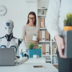 A humanoid robot works on a laptop at a desk while a person carries a box of belongings in an office setting.