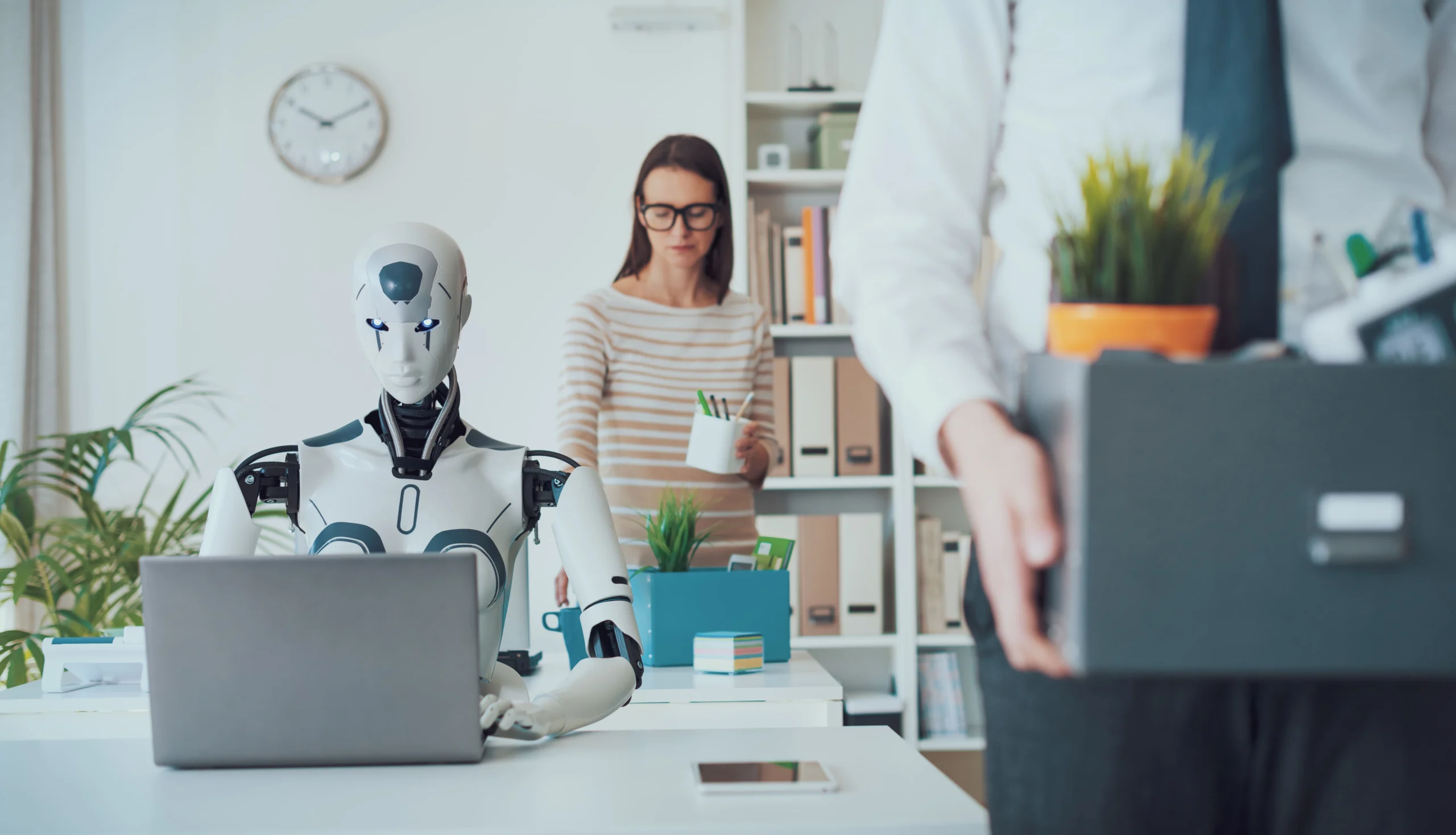 A humanoid robot works on a laptop at a desk while a person carries a box of belongings in an office setting.